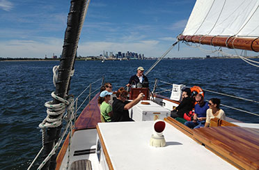 Group of people sitting in the cockpit of Schooner Adirondack III on a Boston Day Sail with the Boston Skyline in the background