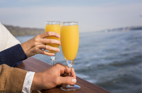 Close up photo of two hands holding mimosas on the bow of the Yacht Northern Lights for a Boston Harbor Fall Foliage Brunch Cruise