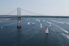 aerial photo of multiple sailboats racing under a bridge for the start of a sailing race