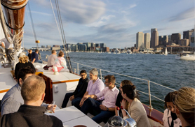 Group of guests sitting in the cockpit of Schooner Adirondack III with the Boston Skyline in the background on a sunny day.