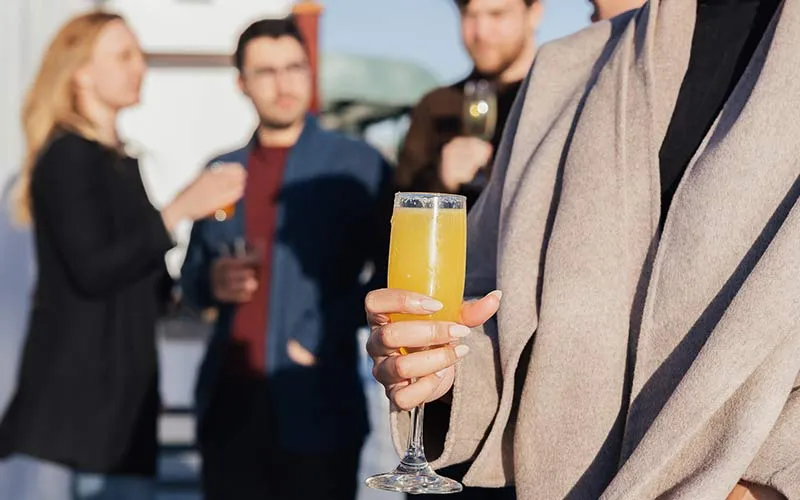 a closeup of a woman holding a glass of mimosa, with a group of people socializing behind her
