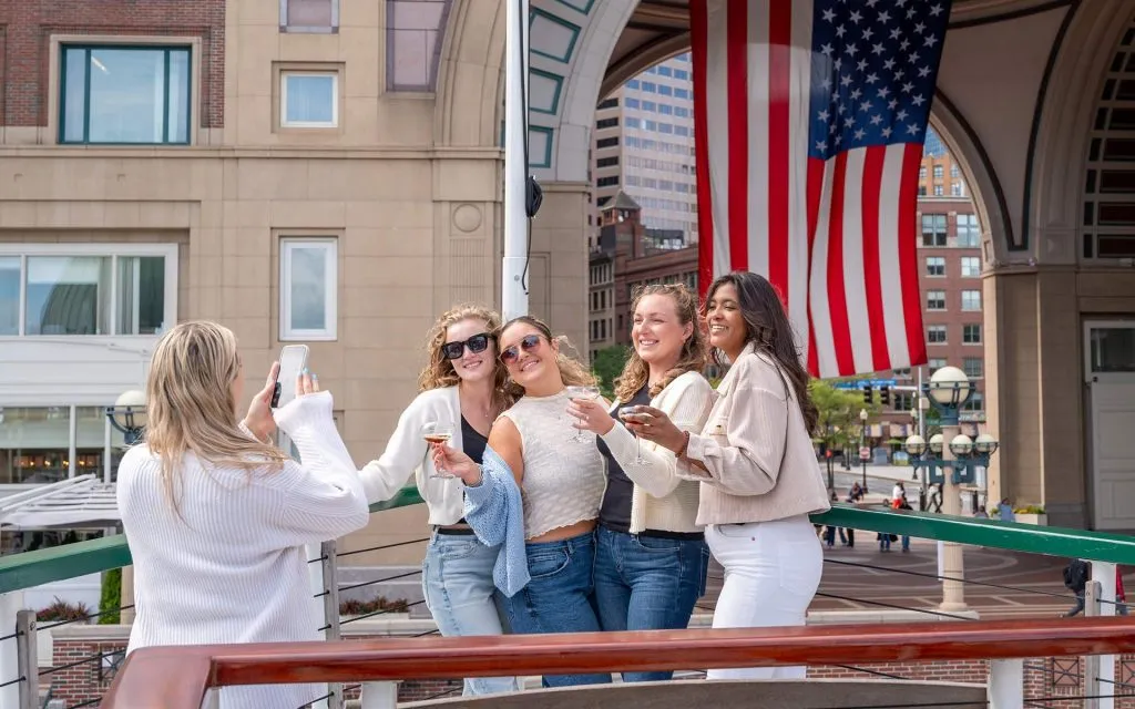 a group of women smiling and posing for a photo, with another woman using her phone for the photo