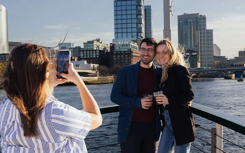 a man and a woman getting their photo taken by someone, aboard a Boston Yacht, with the city in the background