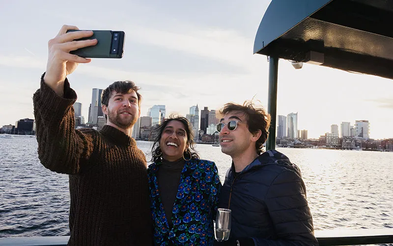 Boston_Northern-Lights-Sunset-Slider-1 two men and a woman taking a selfie together, on a motor yacht, with Boston downtown in the background
