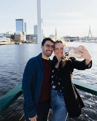 a man and a woman taking a selfie together, at the nose of a motor yacht, with downtown Boston behind them