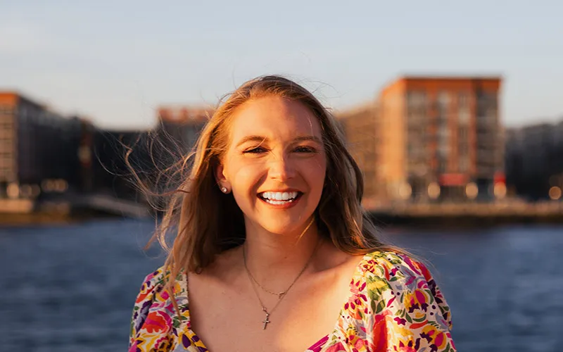 closeup of a woman in a sundress, smiling, with buildings behind her across the water