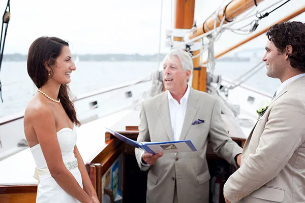 a man and a woman, both dressed in wedding attire, in front of a priest aboard a sailboat