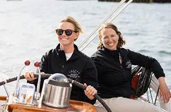 two female sailboat crew members smiling, while steering a Boston sailboat