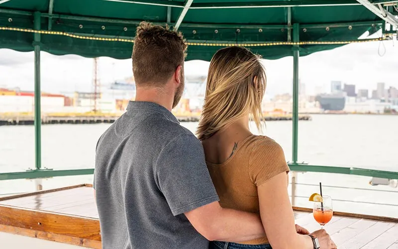 Boston_Our-Mission-Main a man holding a woman around her waist, while both of them look out towards the water aboard a motor yacht