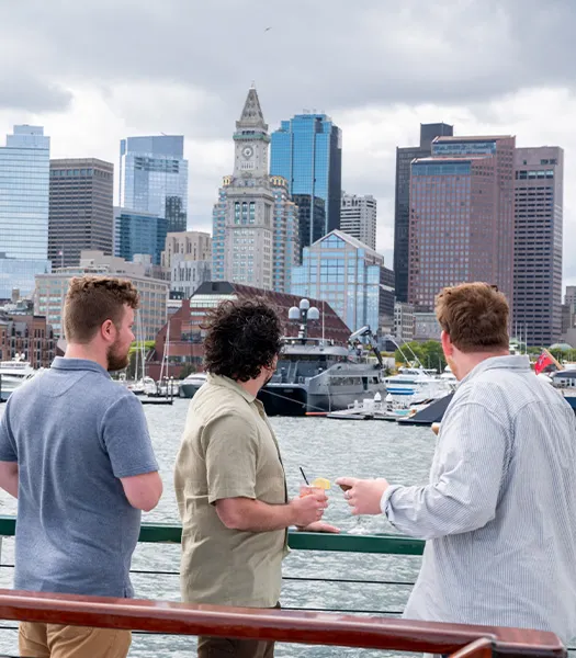 three men, standing at the railing of the motor yacht they are on, looking out towards downtown Boston